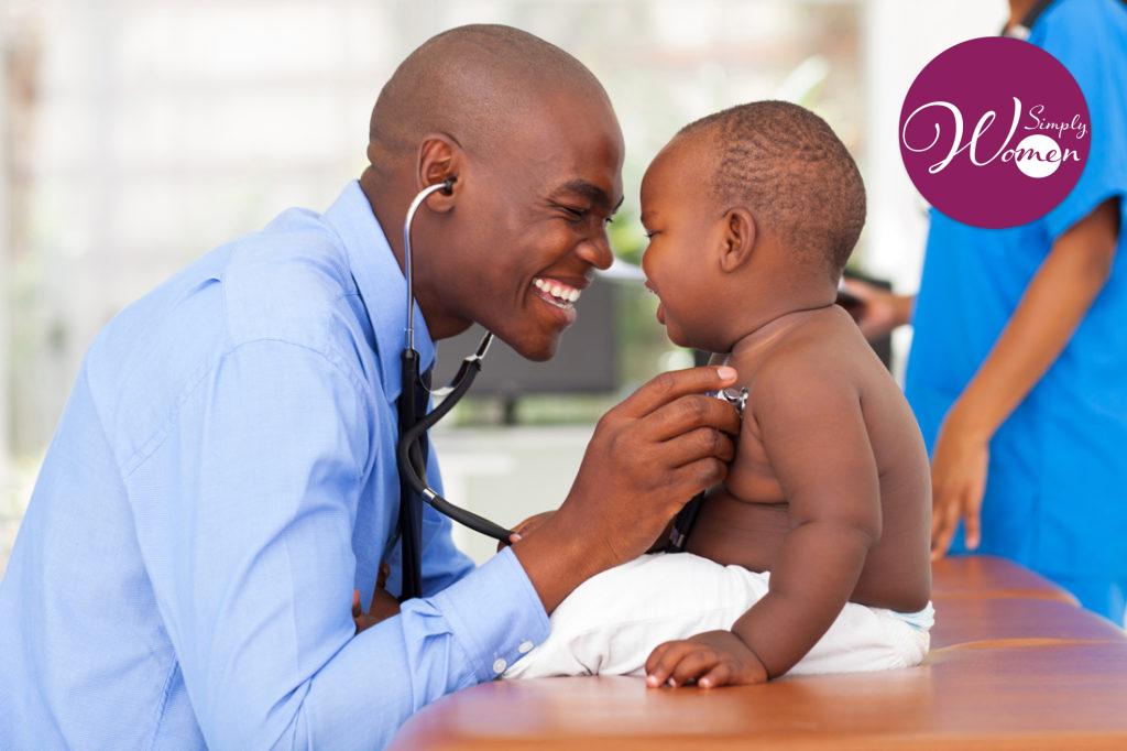 A doctor smiles to elicit a smile from the crying baby he is examining while holding a stethoscope to the baby's chest.
