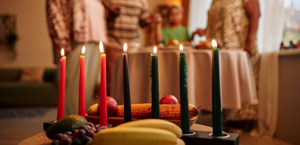 A display of lit candles, fruits and vegetables is in a dining room where the family stands at the table in the background. 