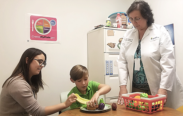 A boy sits at a table with an adult and arranges plastic fruits on a plate while a female caregiver observes.