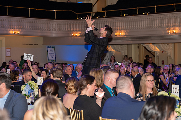 Man in dark suit jacket with microphone, standing on chair with hand raised in crowd during auction
