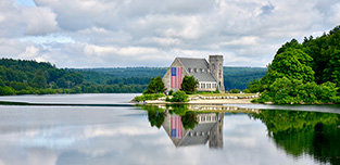 The Old Stone Church in West Boylston is shown next to a lake and cloudy skies.