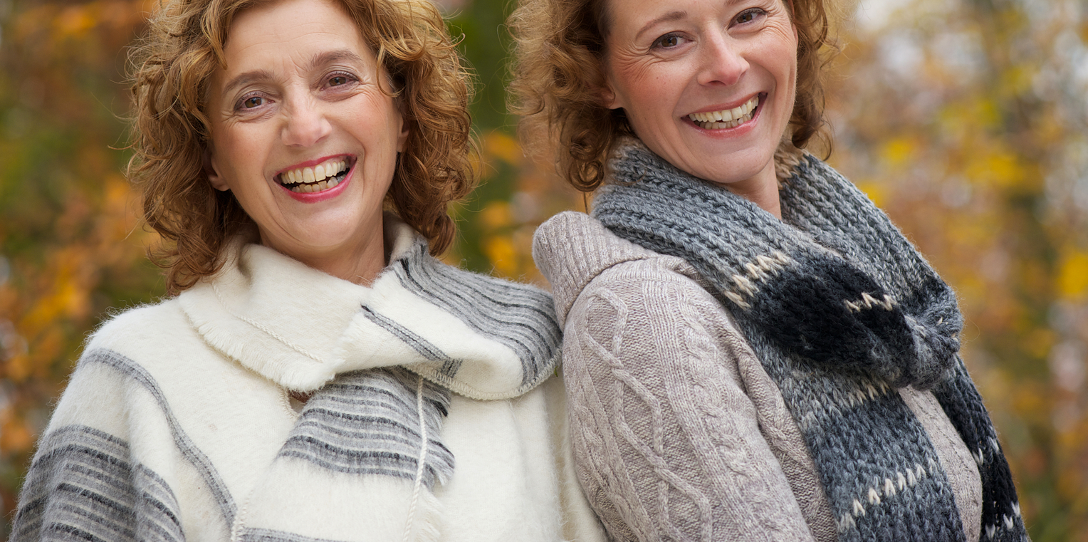 Two women pose for the camera, shoulder-to-shoulder, in a park during autumn