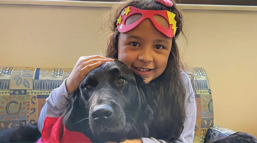 A young patient sits on a sofa, smiling brightly and cuddling Valentina, the black retriever. Valentina is the Child Life Program's animal-assisted therapy dog. 