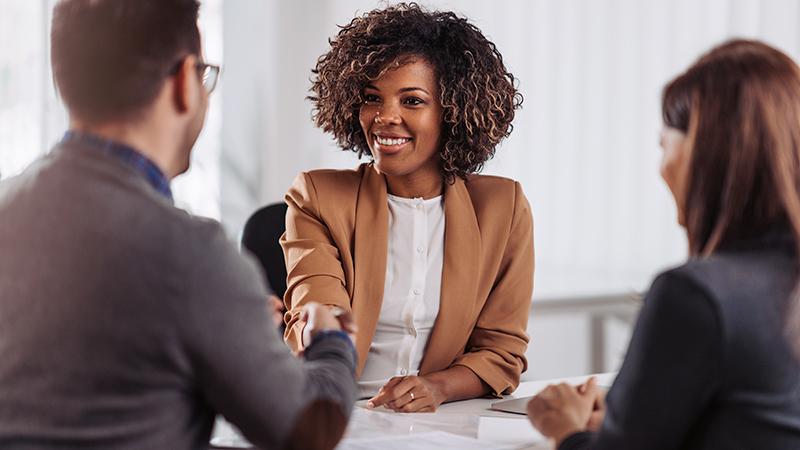 A woman is being greeted with the handshake of one of two job recruiters at a desk during the beginning of her interview.