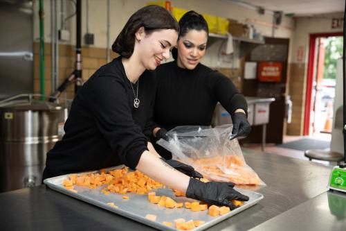 Two women in a hospital kitchen prepare a sheet pan of vegetables.