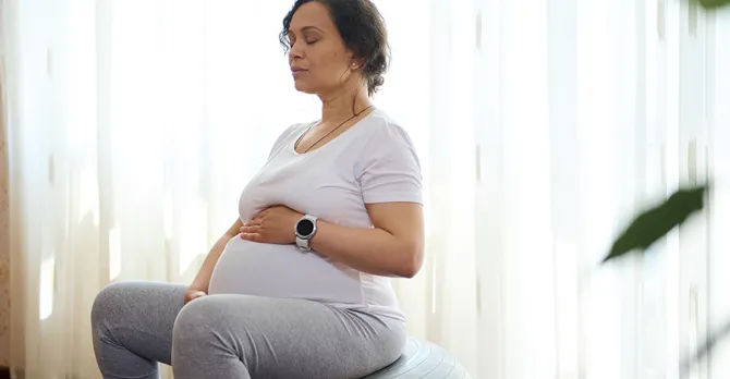 Relaxed conscious pregnant woman holding hands on her belly, doing breathing exercises on a fit ball at home.