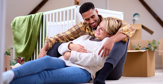An expecting couple sits on the floor of a a room, with a crib, boxes and houseplants behind them.