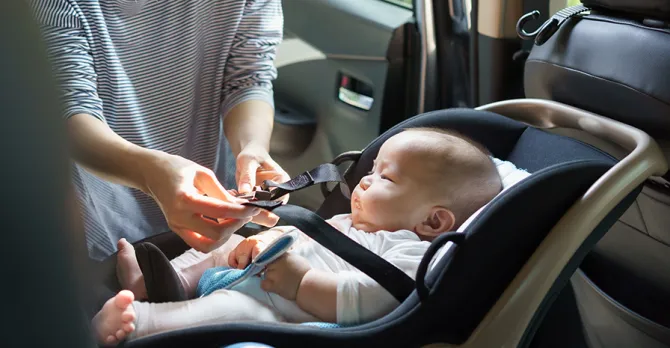 Mother securing her baby in a car seat in the rear passenger side of a car, ensuring safety.