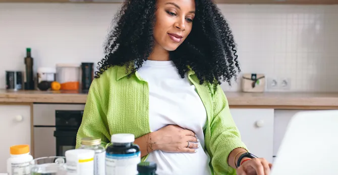 A pregnant woman does research on her laptop, which is next to vitamin bottles.
