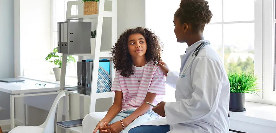A caring, black female doctor talks to her adolescent patient who is sitting on an examination table.