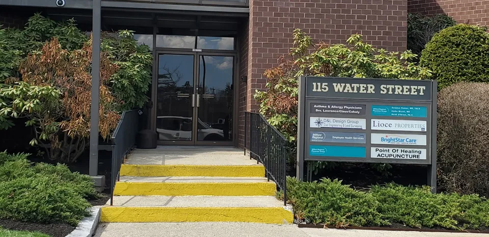 The exterior of a brick building on a sunny day. The steps up are yellow. A sign reads "115 Water Street" and lists the businesses inside.