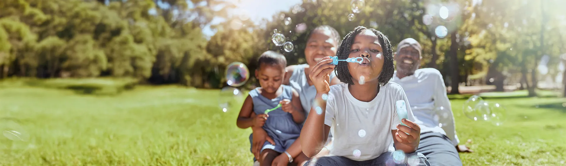 A black family sits on the grass in a park during the summer.  The two children are blowing bubbles.