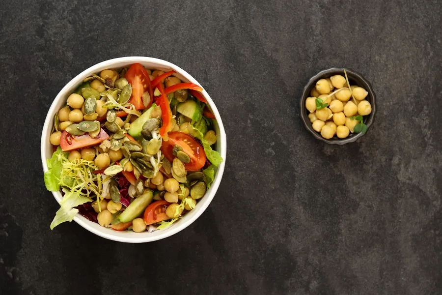 A large white bowl with chickpeas and other Mediterranean vegetables is next to a small bowl of chickpeas, both on a black background.