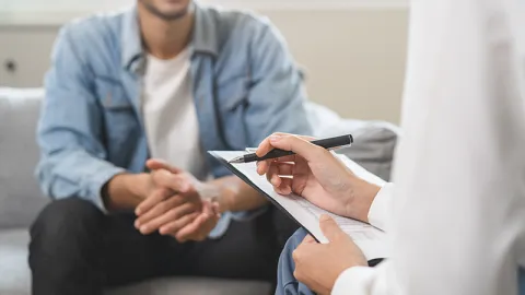 A young man with his hands clasped sitting across from a woman in a white lab coat with a clipboard and pen