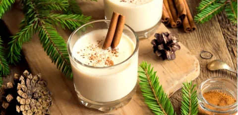cup of eggnog on a wooden cutting board with pinecones and greenery