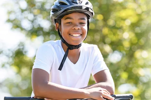 Outdoors on a sunny day, a boy wears his helmet and poses with his forearms across his bicycle handlebars.