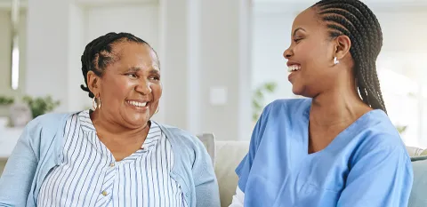 A nurse is sitting on a sofa in a living room with a patient.  Both are laughing.