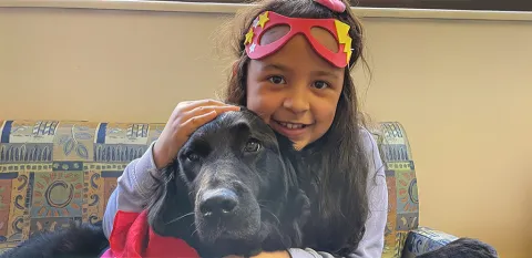 A young patient sits on a sofa, smiling brightly and cuddling Valentina, the black retriever. Valentina is the Child Life Program's animal-assisted therapy dog. 