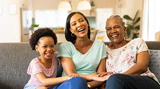 Three generations of females sit together on a sofa and share a laugh: mother, daughter, and grandmother.
