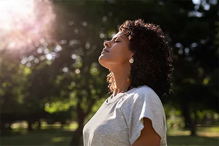 A woman stands with her head tilted back, being mindful and admiring the sun shining through the trees above her.