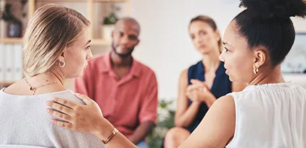 A group of people sits in a circle. A person on the right puts their hand on the shoulder of the person on the left.