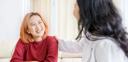A teenager sits with an adult. The teenager is smiling at the adult while the adult has their arm on the teen's shoulder.