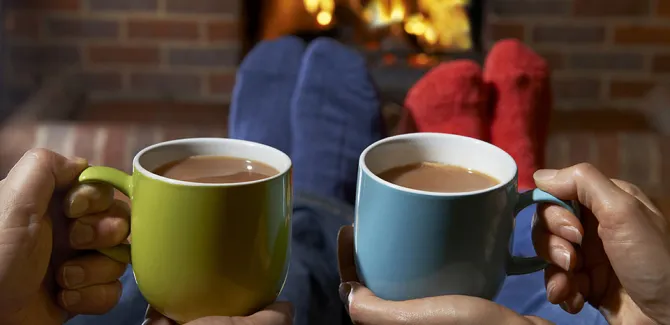 The hands of two people sitting side by side are holding cups of hot chocolate.  The fireplace is just beyond their feet.