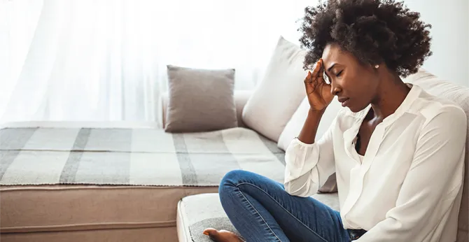 I black woman sitting cross-legged in a chair, hand on forehead and eyes closed.