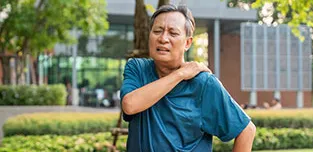 A senior Asian man grimaces, showing he's in pain.  His right arm crosses his chest to place his hand on his left shoulder.