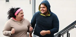 A young woman and man are outside jogging in their sweats on a city sidewalk.