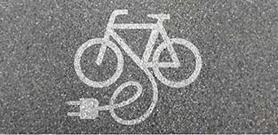 The white painted stencil of of a bicycle and electrical plug connected by a cord painted on pavement, indicating an electric bike charging, placed on black pavement.