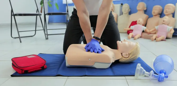 An instructor demonstrates CPR on a test dummy. There is a first aid kit to the left.