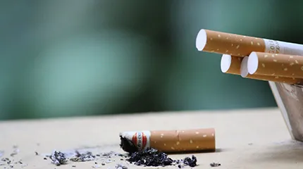 A used cigarette butt and its ashes lies on a surface. To the right, there are four unsmoked cigarettes hanging over the edge of a container.