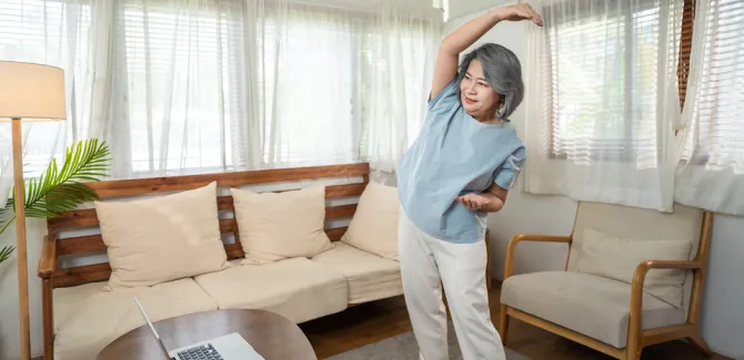 A woman stands in a living room while stretching one arm over her head. She is looking at a laptop on a table while stretching.