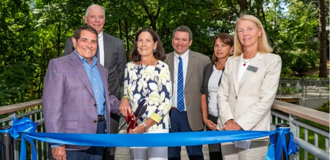 Six people stand in front of a blur ribbon for a ribbon-cutting. They are all looking at the camera, smiling.