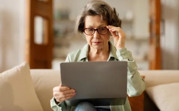 older woman with cataracts wearing glasses struggling to look at a computer.
