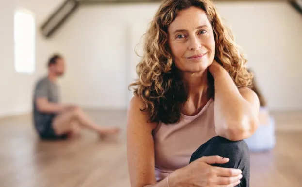 A woman with long hair smiles while sitting on the floor of a yoga, one knee to her chest, one hand behind her neck.