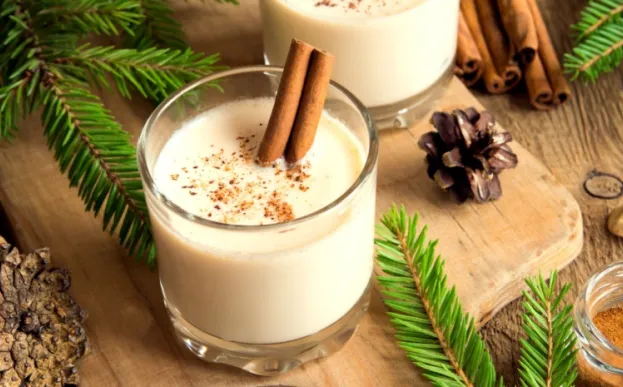 cup of eggnog on a wooden cutting board with pinecones and greenery