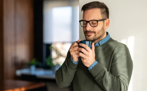 Man with eyes closed holding a warm cup of coffee in a kitchen practicing mindfulness