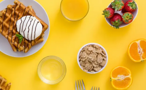 Waffles, cereal, fruit, and juice are shown from above on a yellow table.