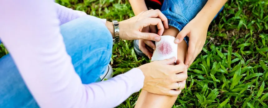 A woman's hands are holding some bandaging over a child's bloody knee.