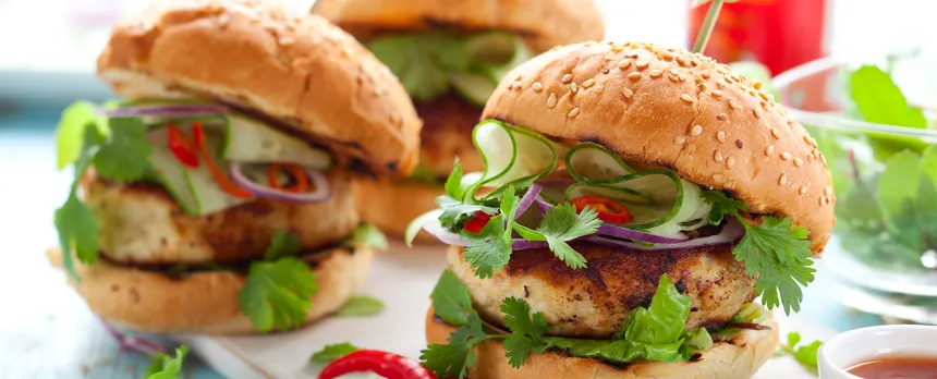 Several turkey burgers with various vegetables on sesame seed buns are shown on a cutting board.