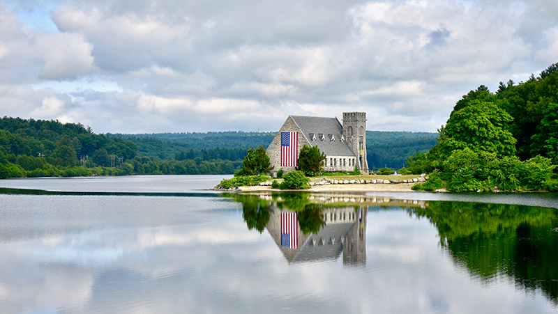 The Old Stone Church in West Boylston is shown next to a lake and cloudy skies.