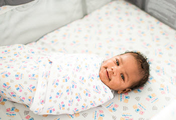 A happy, swaddled baby is shown resting in a crib