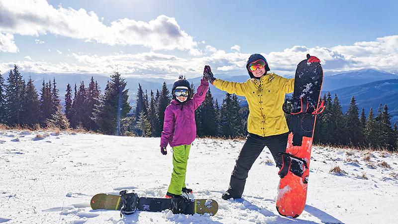 Mother and daughter high fiving at the top of a mountain with snowboards nearby